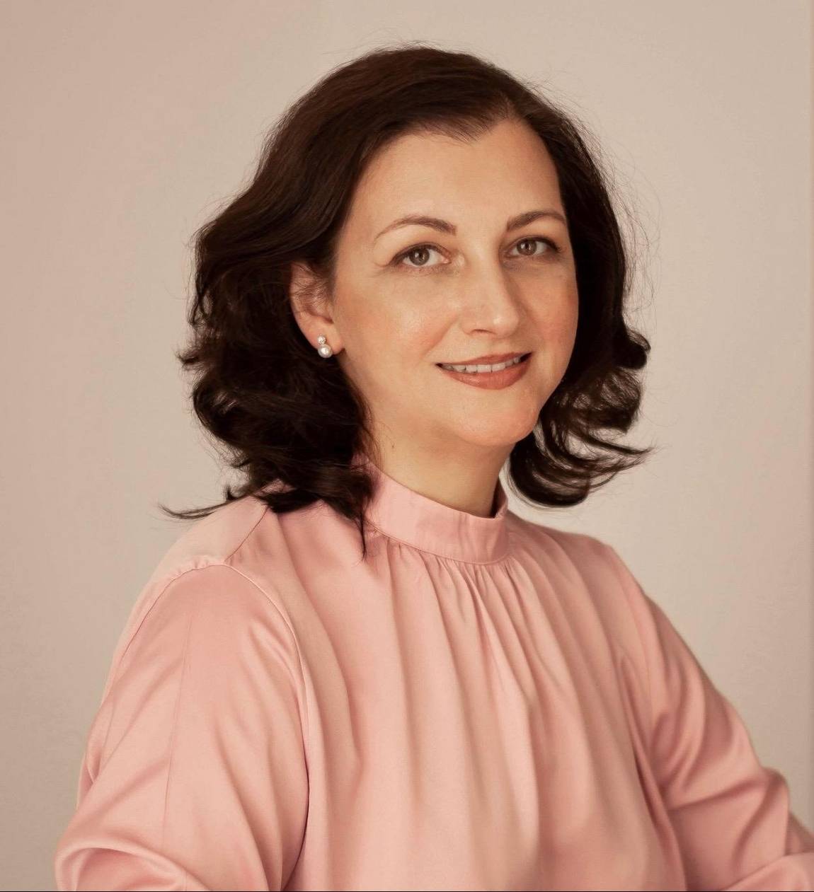 Eine Frau mit schulterlangem dunklem Haar und heller Haut, die eine langärmelige rosa Bluse trägt, lächelt sanft und posiert vor einem schlichten, hellen Hintergrund.